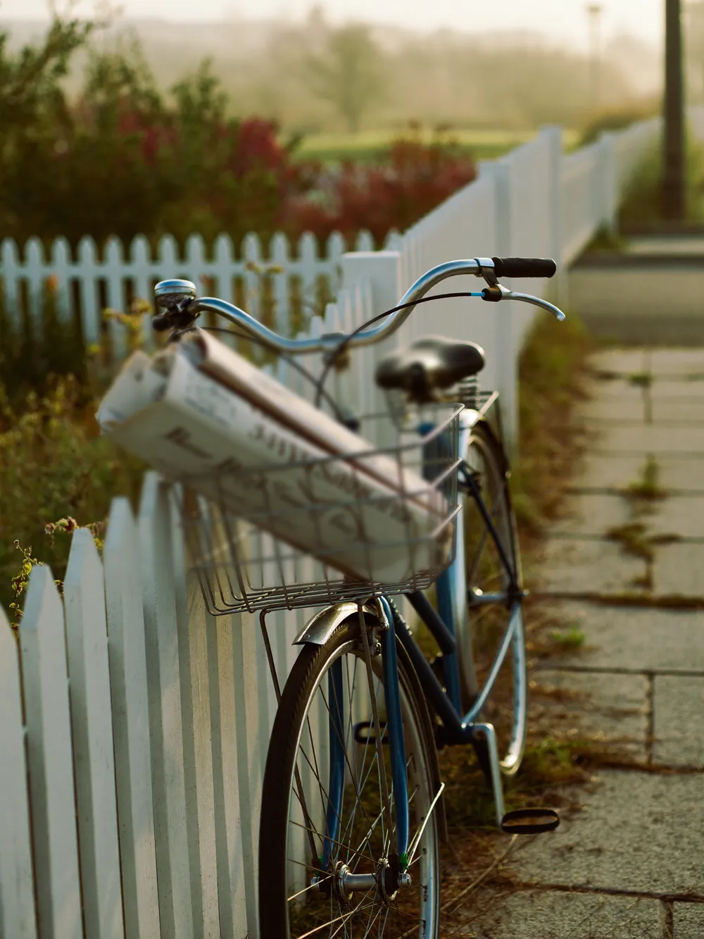 A bicycle with a basket of rolled newspapers leaning on a white picket fence in early morning light